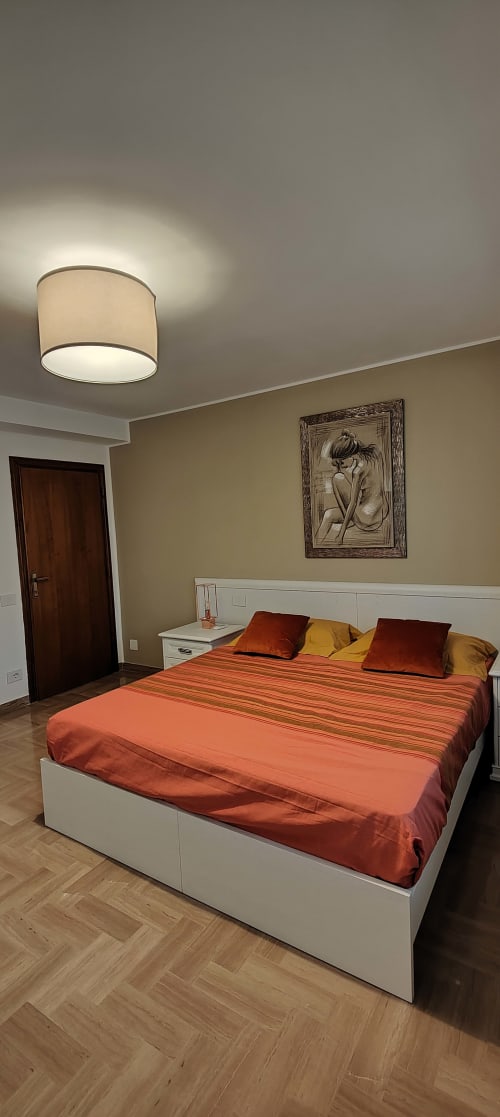 Camera da letto moderna con letto bianco, biancheria arancione, plafoniera a soffitto, quadri incorniciati e pavimentazione in legno nello stile italiano