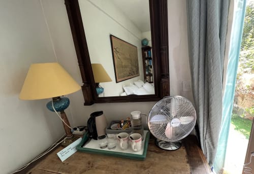 Coin chambre douillet à la Maison 1850 Paris avec lampe de bureau, ventilateur, miroir et vue sur le jardin par la fenêtre drapée