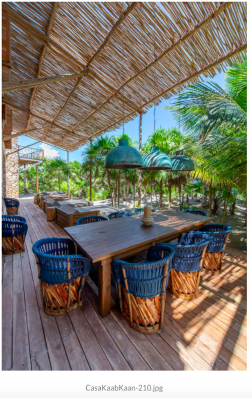 Comedor de villa frente al mar en Tulum con mesa de madera, sillas tejidas azules y techo de palapa con vista a palmeras