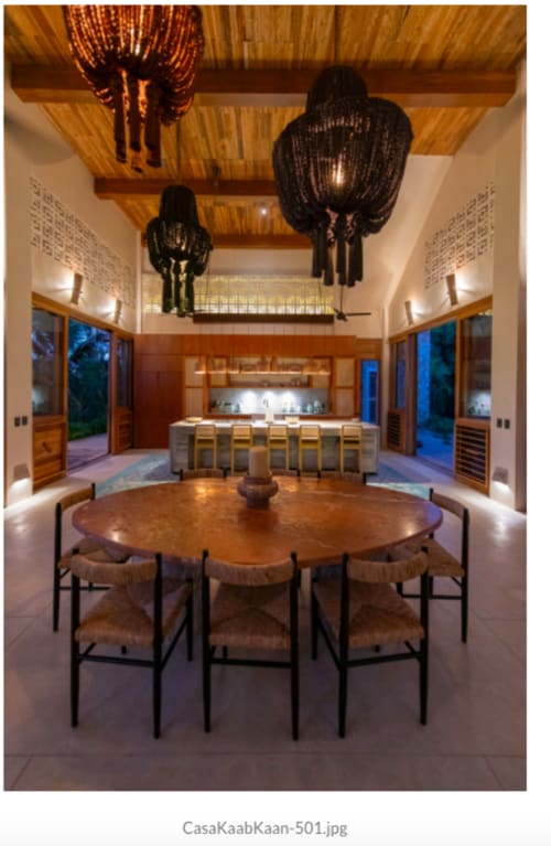 Comedor de villa moderna en Tulum con mesa redonda de madera, lámparas colgantes negras, cocina al aire libre y vistas al mar a través del cristal