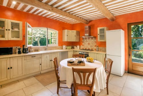 Une cuisine lumineuse avec des murs orange, des poutres en bois et une table à manger ronde au Mas des Olivades, une villa à Pernes-les-Fontaines, en Provence, France.