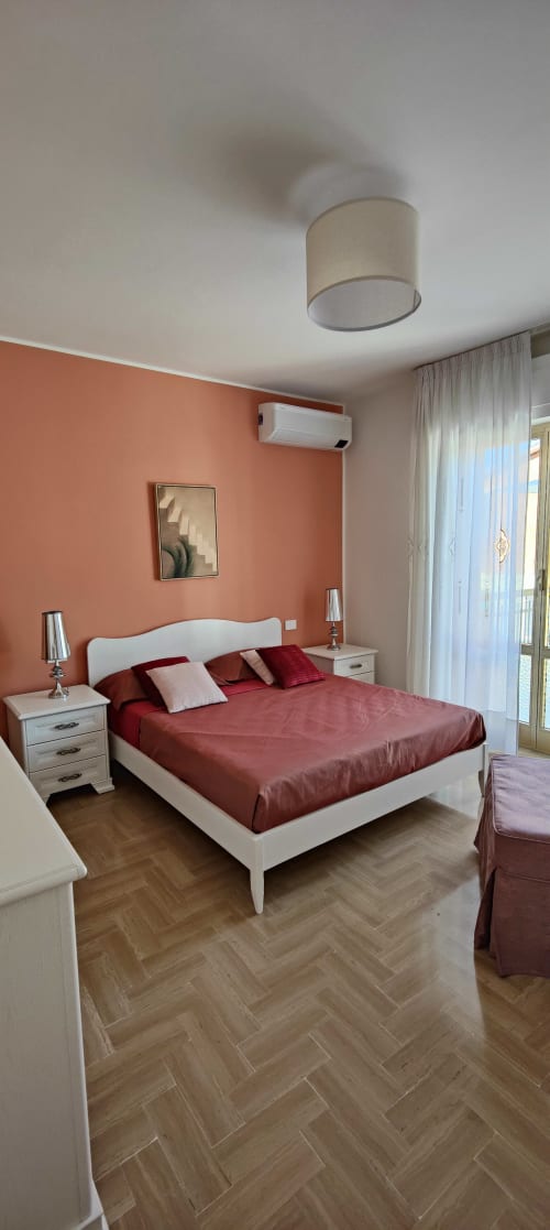 Modern bedroom with coral accent wall, white bed, burgundy bedding, and lake views at Villa Italia