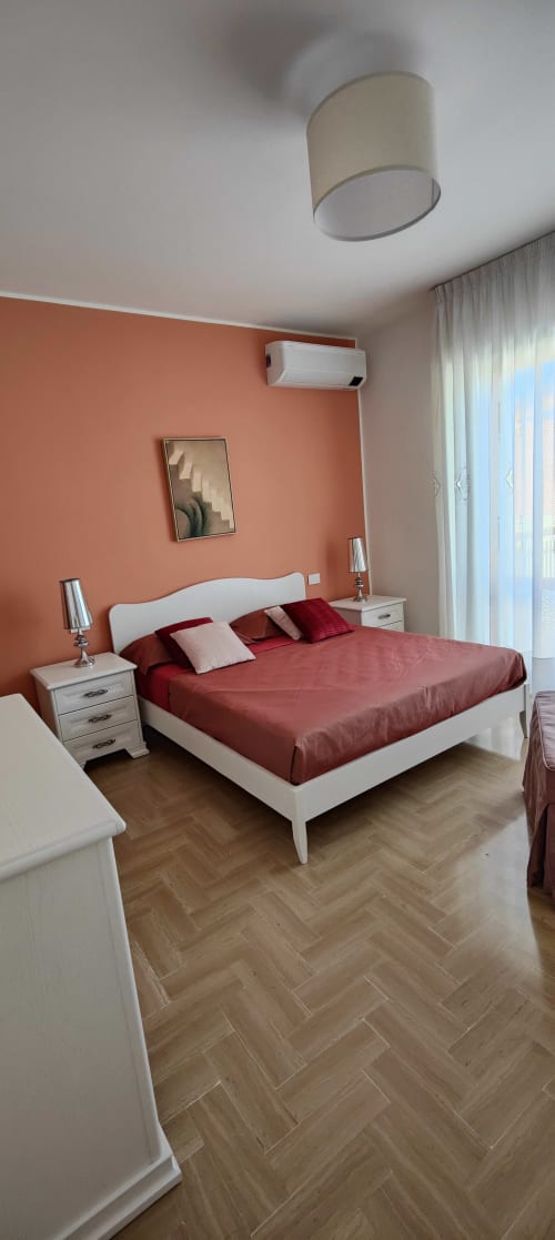 Modern bedroom at Villa Italia with coral accent wall, white bed, burgundy linens, and lake view through curtained window