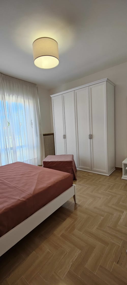 Modern bedroom with red bedding, white wardrobe, cylinder ceiling light, wood flooring, and sheer curtains at Villa Italia