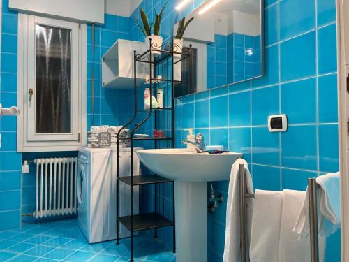 Bagno in piastrelle azzurre luminose nell'Apartment GioviGemini, Treviso con lavabo su colonna, asciugamani bianchi e moderna unità di scaffali neri