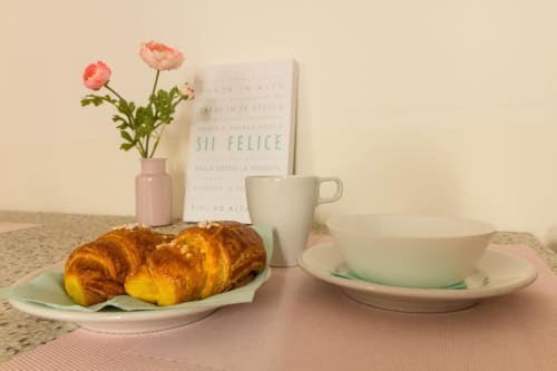 Colazione agli Appartamenti Ferrara: pasticcini, tazza di caffè, fiori rosa freschi e cartolina italiana su terrazzo