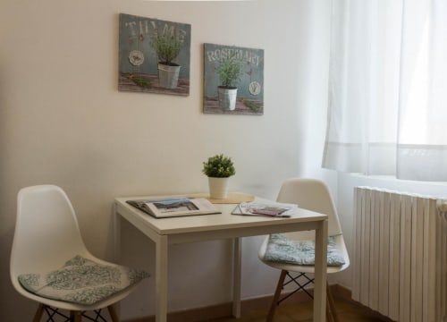 Luminoso angolo di appartamento con tavolo da pranzo bianco, due sedie, piante in vaso e arte murale a tema erbe presso Appartamenti Ferrara
