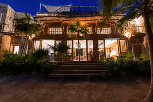 Villa Kaab Kaan en Tulum con fachada frente al mar iluminada, pérgola de madera, plantas de palma y vistas al mar al atardecer
