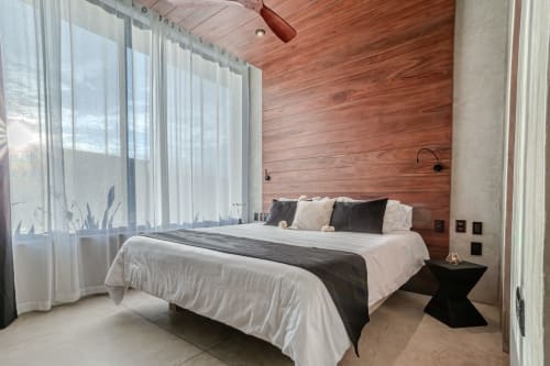 Dormitorio de villa moderna en Tulum con pared de acentos de madera, ropa de cama blanca, cortinas translúcidas y vistas al jardín desde ventanales