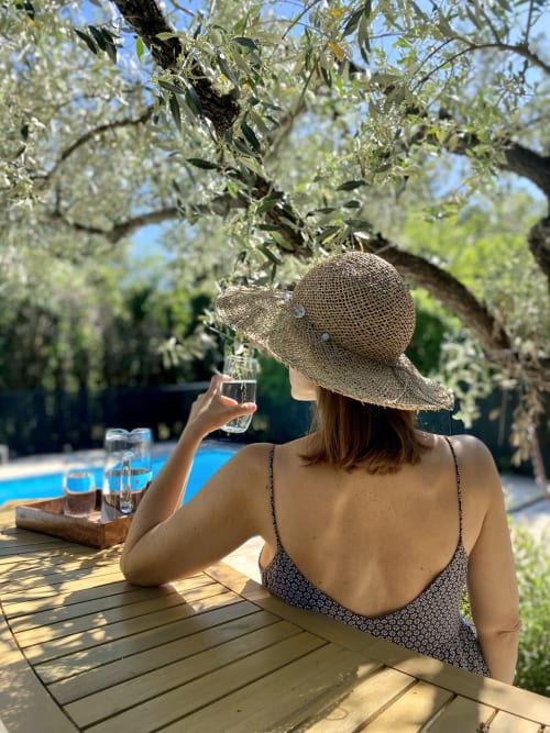 Une femme portant un chapeau de paille et tenant un verre est assise à une table avec vue sur la piscine au Mas des Olivades, une villa à Pernes-les-Fontaines, en Provence, France.
