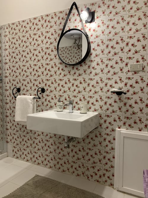 Bagno moderno dell'Agriturismo Il Pomo d'oro con carta da parati floreale, specchio rotondo, lavabo bianco e rubinetteria nera ad Assemini