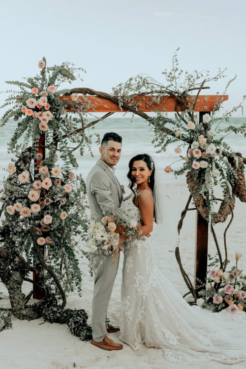 Couple in wedding attire under floral arch on beach in Tulum, Mexico