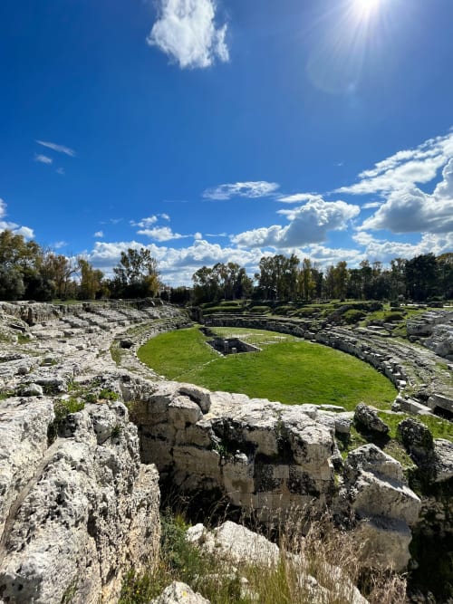 Rovine dell'antico teatro greco a Siracusa con arena verde e gradinate in pietra, cielo blu con nuvole bianche