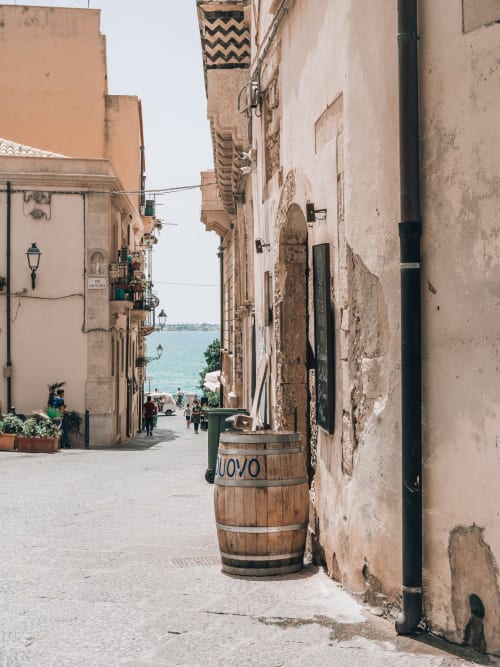 Stretto vicolo in pietra a Siracusa che conduce al mare turchese, edifici dall'aspetto antico, botte di legno, caratteristica via costiera storica