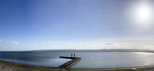 Two people walking on pier at West Kirby beach with calm water and distant hills under blue sky