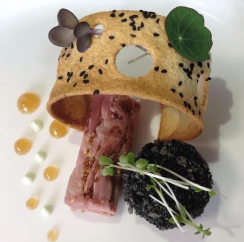 Artistic plated dish at Three Little Pigs hotel featuring sesame seed tuile, cured meat, black garnish and microgreens
