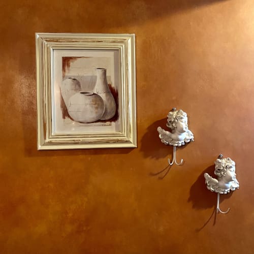 Un tableau de vases sur un mur orange avec deux crochets décoratifs en forme d'ange au Mas des Olivades, une villa à Pernes-les-Fontaines, en Provence, France.