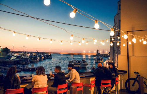 Cena a bordo mare a Bari: luci decorative, sedie rosse e barche nel porto al tramonto