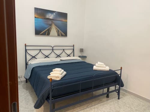 Newhouse apartment bedroom in Bari with metal bed frame, navy bedding, white towels, and seascape artwork
