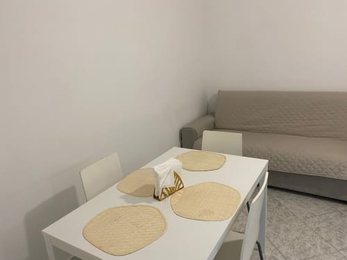 Appartamento Newhouse a Bari: zona pranzo minimalista con tavolo bianco, tovagliette intrecciate e divano grigio sullo sfondo