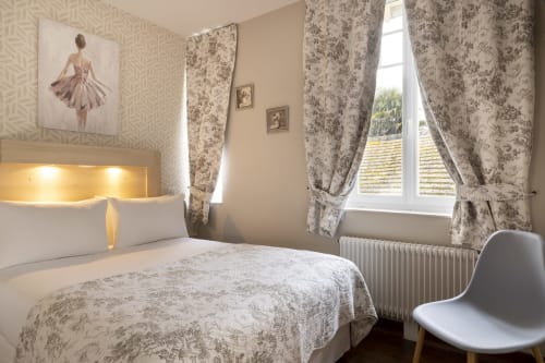Chambre élégante de l'Hôtel De Lion Sur Mer avec rideaux fleuris, tête de lit illuminée et vue balcon sur Lion sur Mer