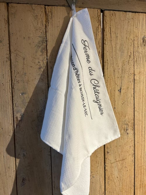 Serviette de cuisine blanche avec texte français suspendue à un mur de bois rustique au gîte La Ferme du Châtaignier près de Paris