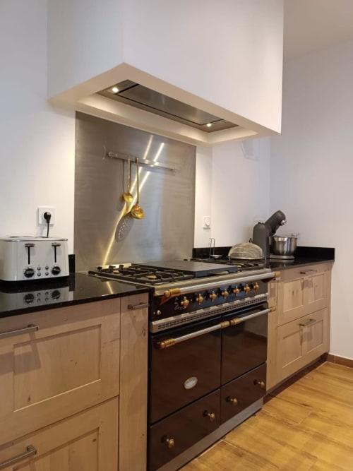 Cuisine moderne avec cuisinière noire, placards en bois clair, crédence inox et hotte aspirante à La Ferme du