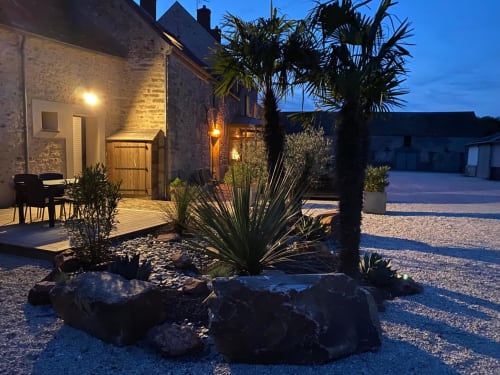 Cour du soir du gîte La Ferme du Châtaignier en Essonne avec bâtiment en pierre éclairé, plantes tropicales et graviers