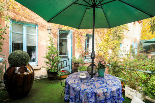 Terrasse de jardin parisien à la Maison 1850 avec parasol vert, table bleue à motifs, murs de briques couverts de lierre et plantes en pot