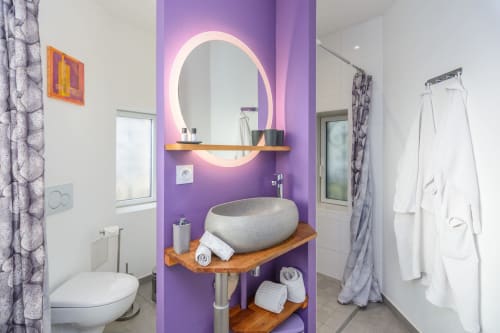 Salle bain de la suite de couleur violette avec à gauche coin toilettes avec rideau au milieu une grande vasque avec miroir grossissant et à droite une grande douche