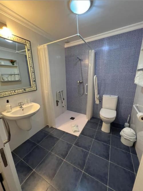 Modern accessible bathroom with dark tile flooring, walk-in shower, toilet, sink with mirror, and blue textured walls at