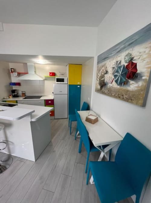 Cocina-comedor moderno con muebles blancos, sillas azules, unidades de almacenamiento coloridas y arte de sombrilla de playa en Complejo