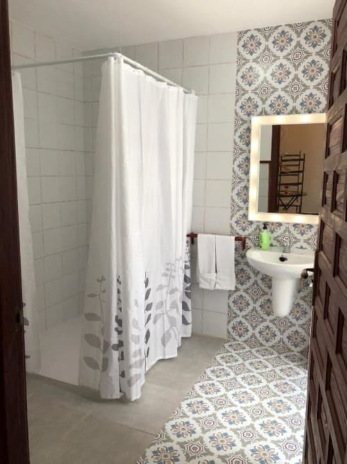Modern bathroom with white tile walls, decorative patterned floor, pedestal sink, lighted mirror, and shower with white