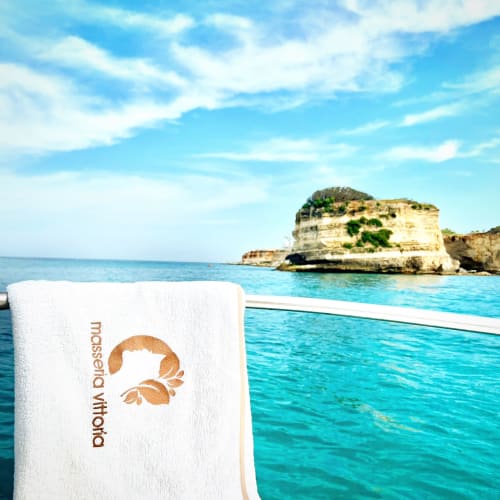 White towel with Masseria Vittoria logo overlooking turquoise sea and rocky island in Acaya, Italy