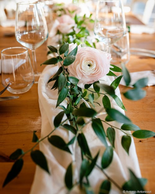 Elegante mise en place a Masseria Vittoria con fiori di ranuncolo rosa, ghirlanda di verde e calici di vino su tavolo di legno
