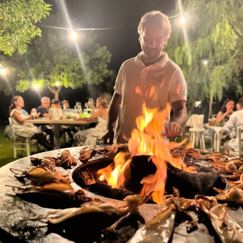 Man tending bonfire at Masseria Vittoria outdoor gathering in Acaya, guests dining under evening lights