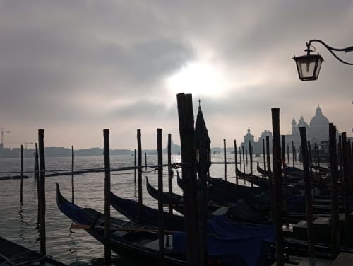 Gondole ormeggiate al fronte lagunare di Venezia con basiliche storiche silhouettate contro il cielo nuvoloso