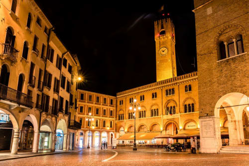 Piazza medievale illuminata a Treviso con edifici storici e torre dell'orologio di notte