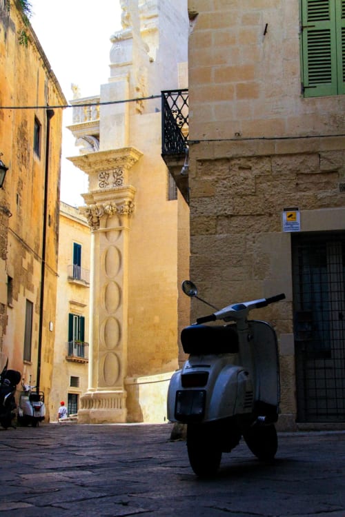Vespa stationnée sur la place du village historique d'Acaya avec architecture baroque ornementée