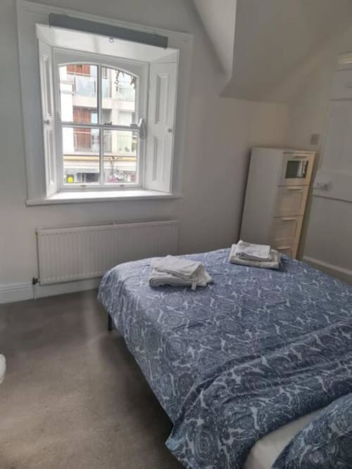 Bright bedroom in Dublin cottage with blue patterned bedspread, open window with city view, radiator, and wooden wardrobe