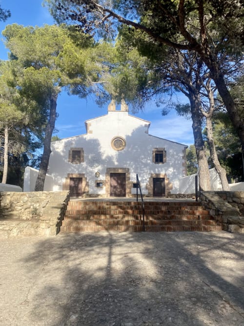 Capilla histórica blanca en BondBeach Village de Altafulla, enmarcada por árboles altos bajo un cielo azul despejado