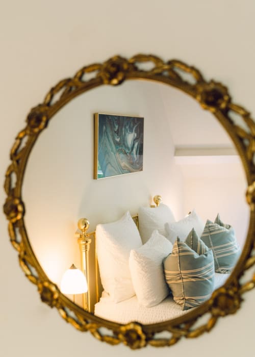 Espejo ornamentado con marco dorado reflejando un elegante dormitorio con almohadas blancas y cojines a rayas en la casita Domaine de Bize