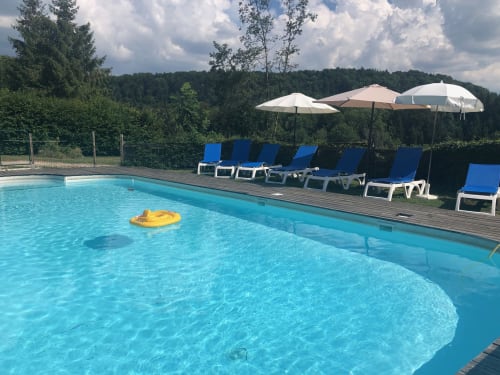 Piscine bleu cristallin avec bouée jaune aux Gîtes Des Planchettes, transats, parasols et vue sur la forêt
