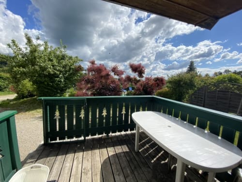 Terrasse couverte des Gîtes Des Planchettes avec table blanche, balustrade verte, vue sur la forêt et ciel bleu