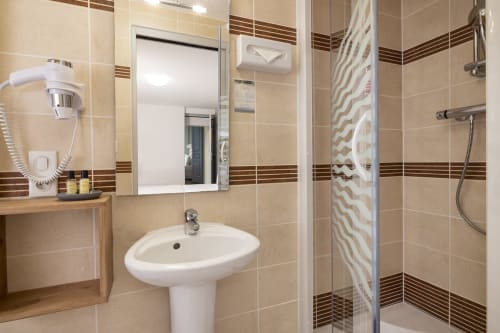 Salle de bain moderne de l'Hôtel De Lion Sur Mer avec carrelage beige, lavabo sur piédestal, miroir et douche à l'italienne avec paroi vitrée