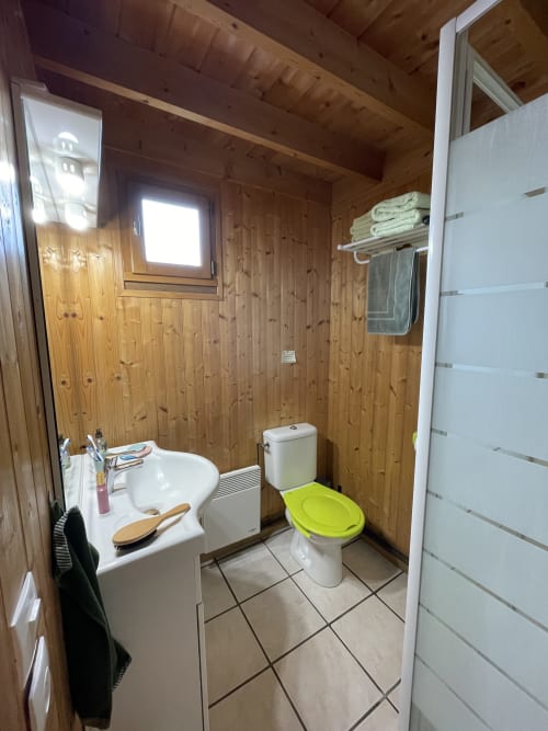 Salle de bain du gîte avec murs en bois, lavabo blanc, toilettes avec siège vert citron, douche, porte-serviettes et fenêtre