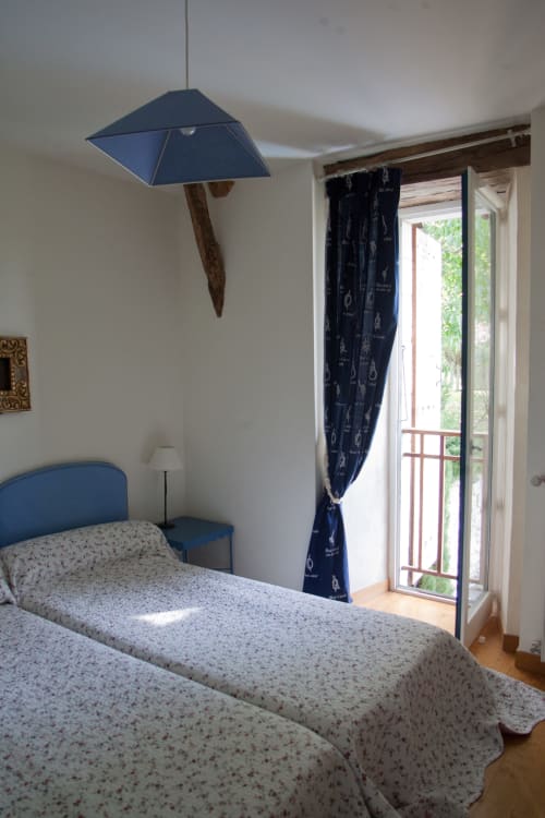 Chambre avec suspension bleue et poutre en bois, couvre-lit fleuri, rideaux foncés et portes vitrées ouvrant sur le jardin