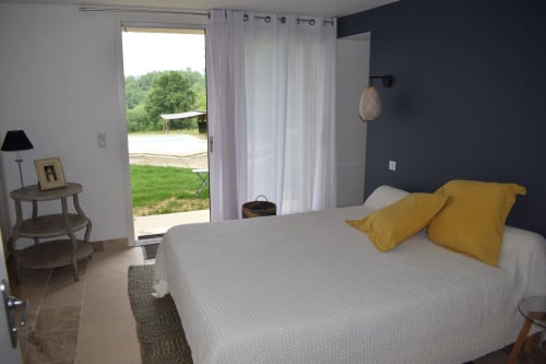 Chambre lumineuse du gîte Les Pratges avec vue sur la piscine, oreillers jaunes sur le lit, mur d'accent sombre, portes vitrées ouvertes sur le jardin