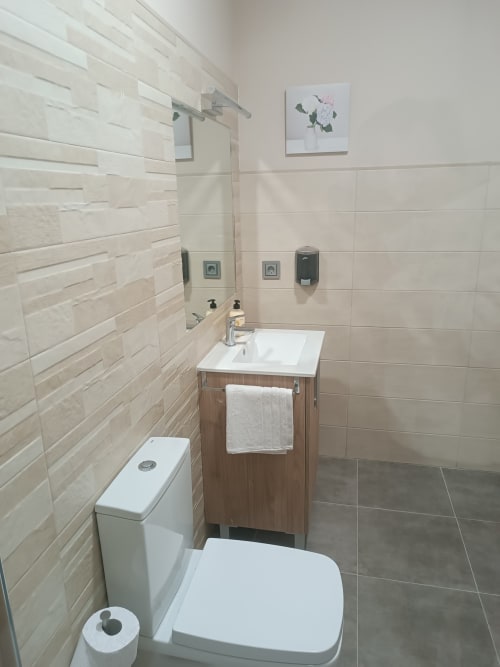 Baño moderno en apartamento de Ávila con paredes de azulejos blancos, tocador de madera, inodoro blanco y arte floral decorativo sobre el lavamanos