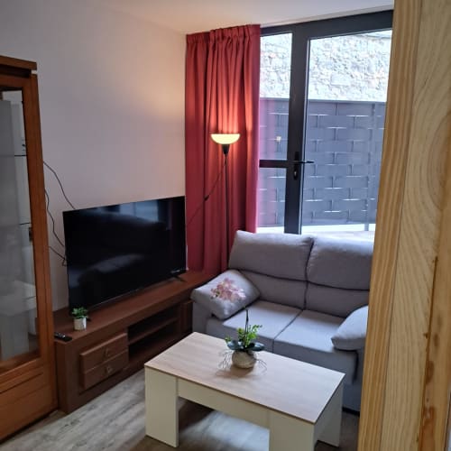 Moderno salón de apartamento en Ávila con sofá gris, televisor de pantalla plana, cortinas rojas y acceso a balcón con vistas a la ciudad.